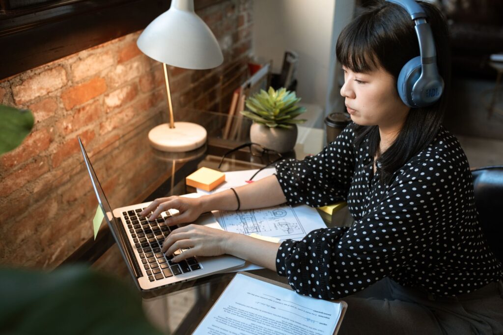 A graduate in headphones typing at a laptop, with printed papers spread across the desk