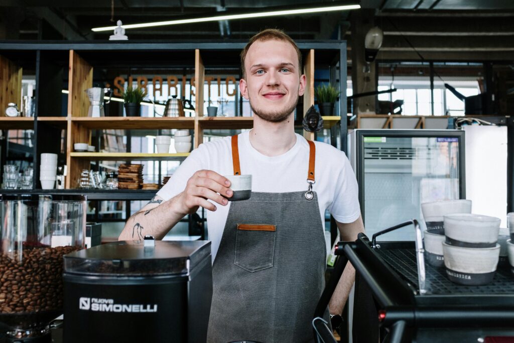 Barista in apron smiling and serving coffee at a modern café. Interior shot with coffee equipment.