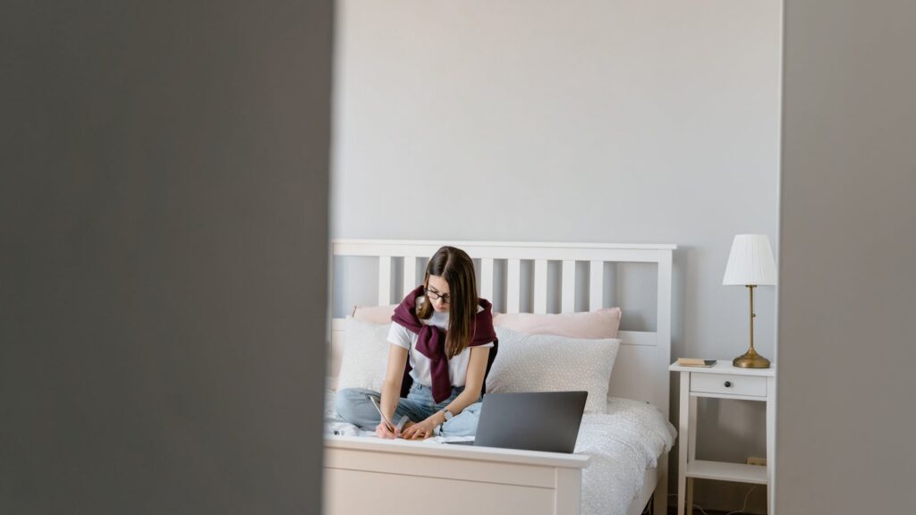 Bright UK student bedroom with laptop on a wooden desk, bed and storage in the background