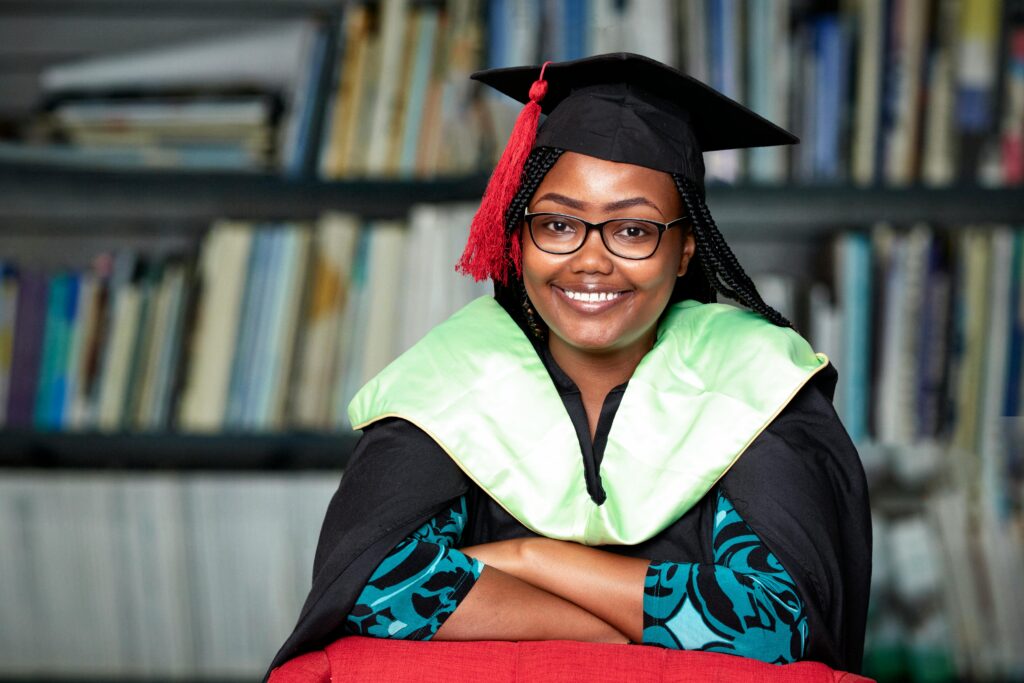 Smiling woman in graduation gown and cap sitting in a library, looking confident.
