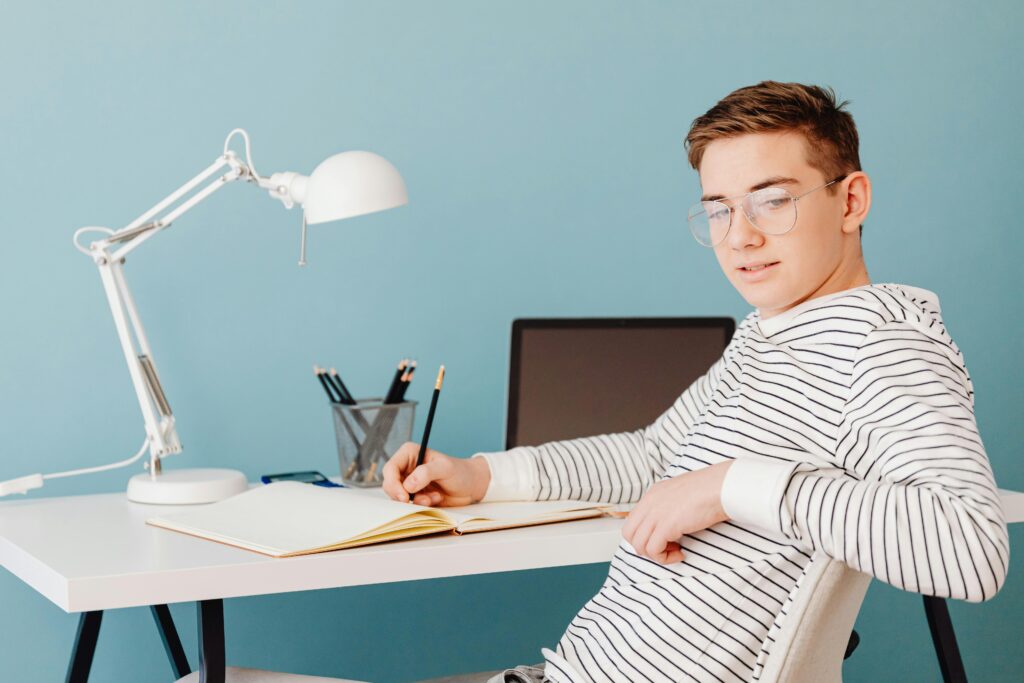 Young male student wearing glasses studying at a desk with a laptop and lamp.