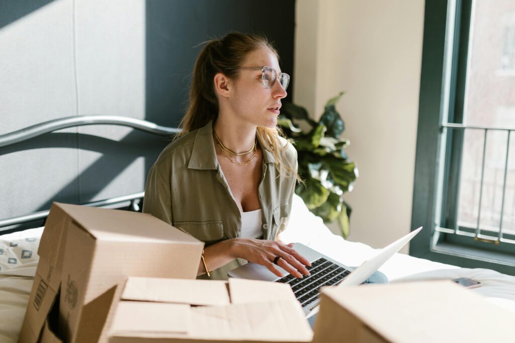 A woman uses a laptop for online shopping surrounded by packed boxes.