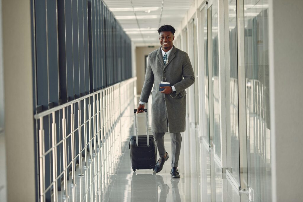 Full length of happy African American male in stylish classy gray clothes with suitcase and passport walking in contempo