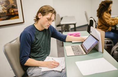 Student writing notes at a desk with laptop
