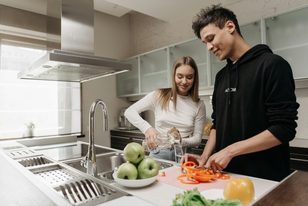 A young couple happily prepares a meal, slicing vegetables and pouring water in a modern kitchen.