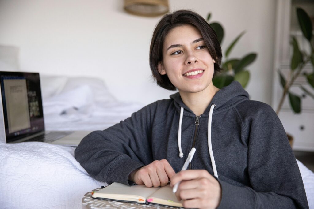 Young woman smiling while writing in a notebook, with a laptop in the bedroom setting.