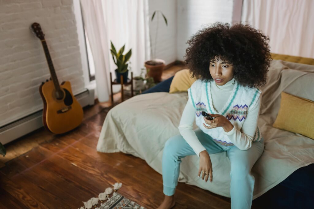 High angle of relaxed young African American female with curly hair switching channels with remote controller while watc