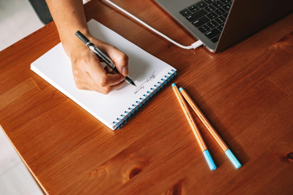 A close-up view of a hand writing notes on a notepad with a laptop nearby on a wooden desk.