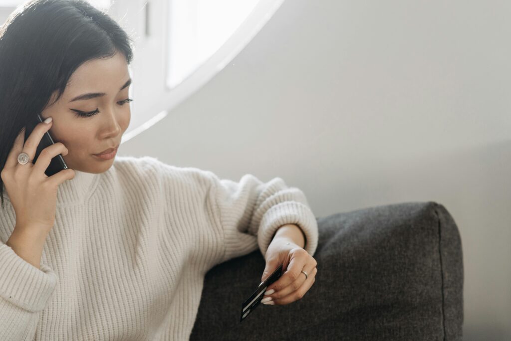 Young woman on phone holding credit card, making an online purchase comfortably at home.