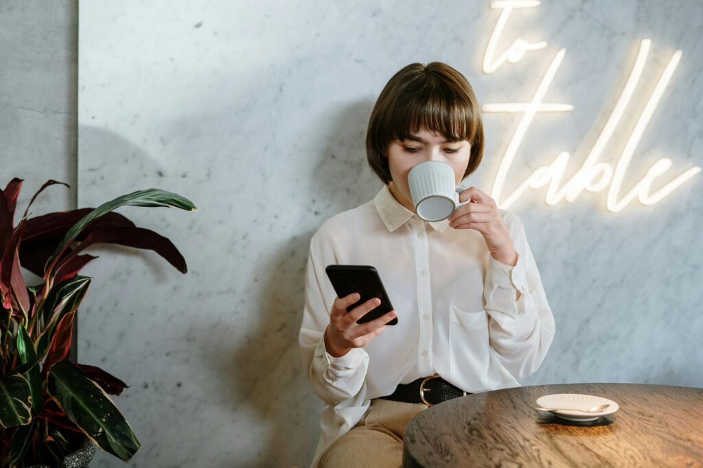 Stylish café scene with young woman enjoying her coffee, using a mobile phone under neon light ambiance.