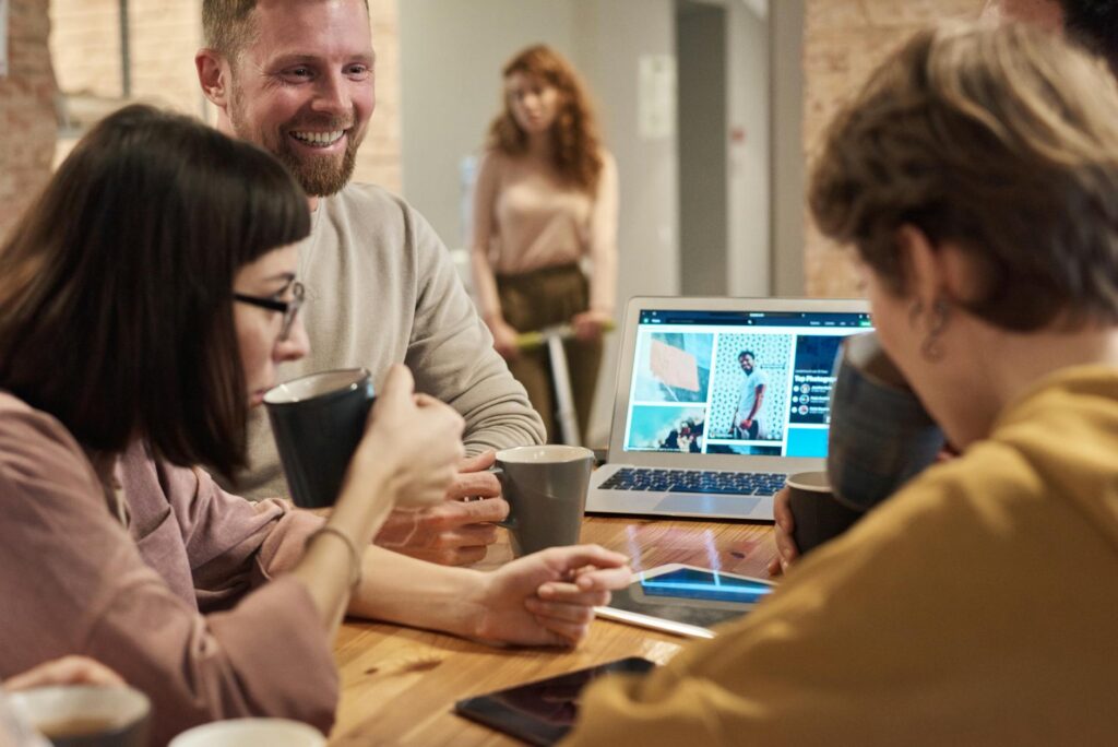 Smiling colleagues with coffee mugs gathered around a laptop in a brick-walled office