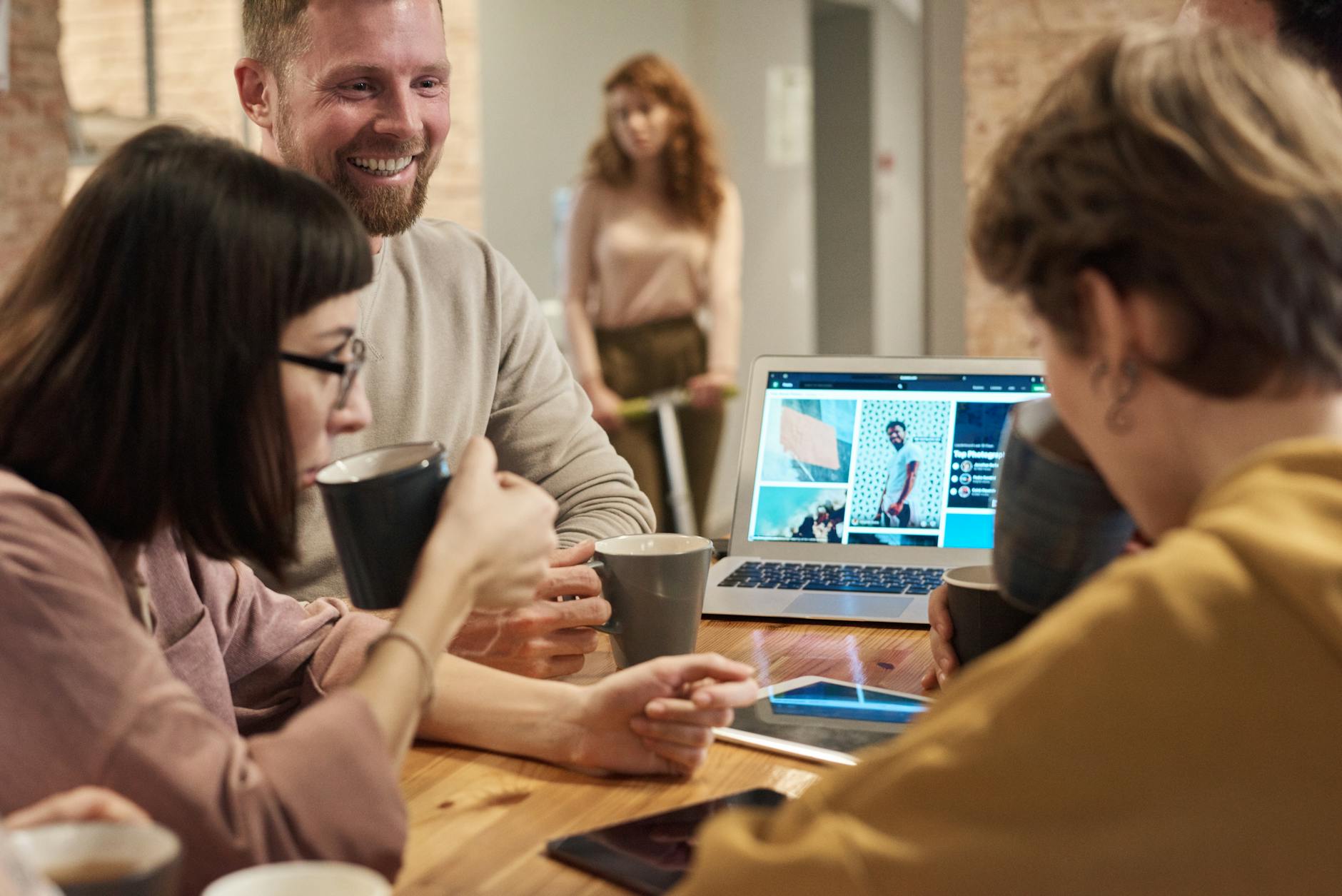 Smiling colleagues with coffee mugs gathered around a laptop in a brick-walled office