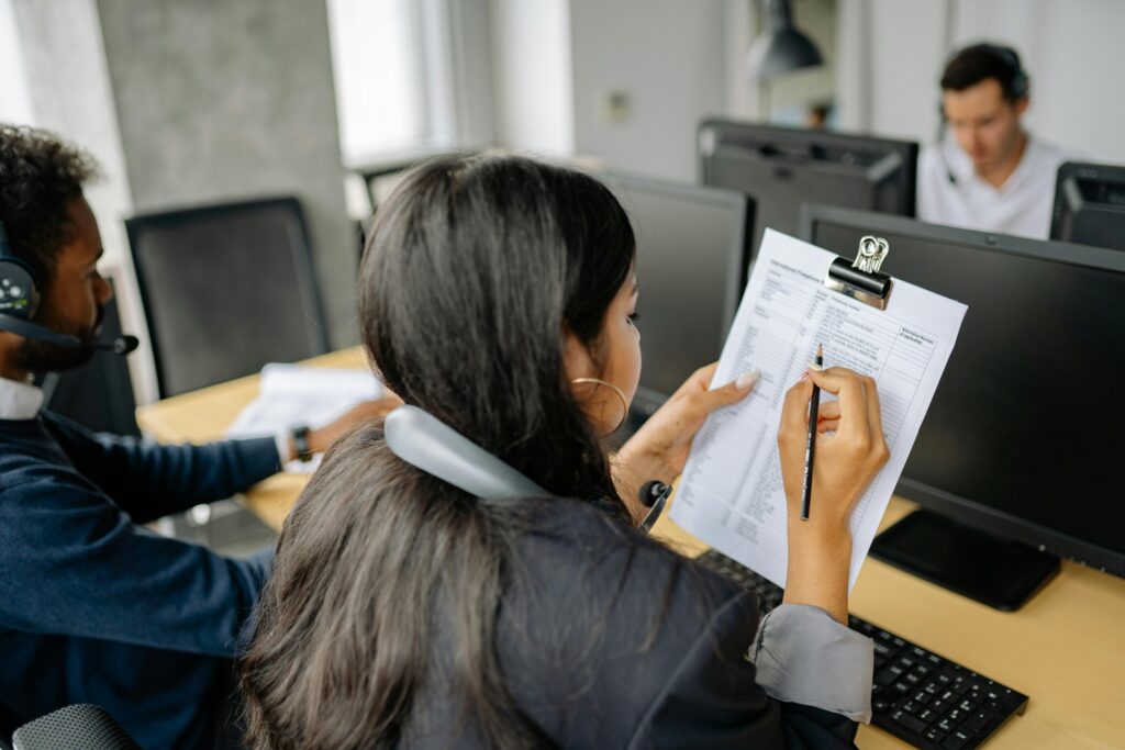 Young staff in headsets doing call-centre jobs, one checking a contact list on a clipboard