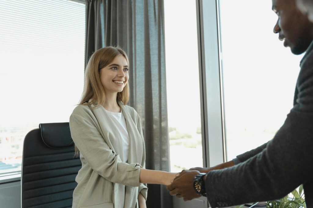 A smiling woman shakes hands with an interviewer across a desk in a bright office