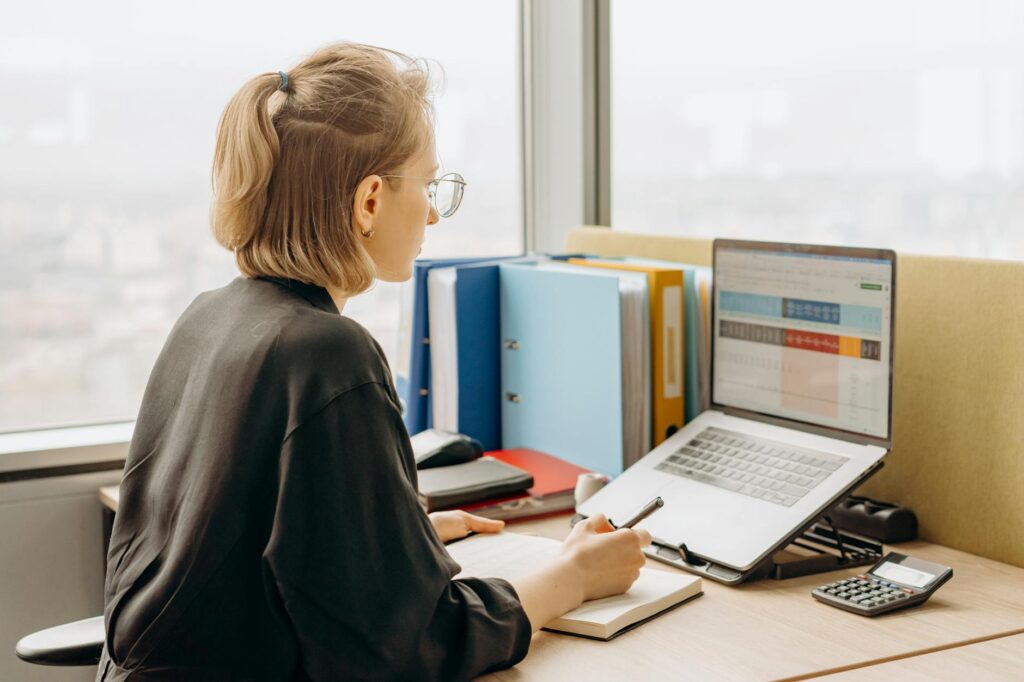 Young professional with a notebook and calculator planning out figures on a laptop spreadsheet