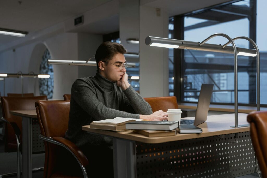 A young man works on a laptop at a desk in a quiet modern university library