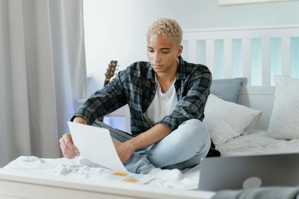 Young man on a bed with an open laptop and notebook, focused on the screen during home study