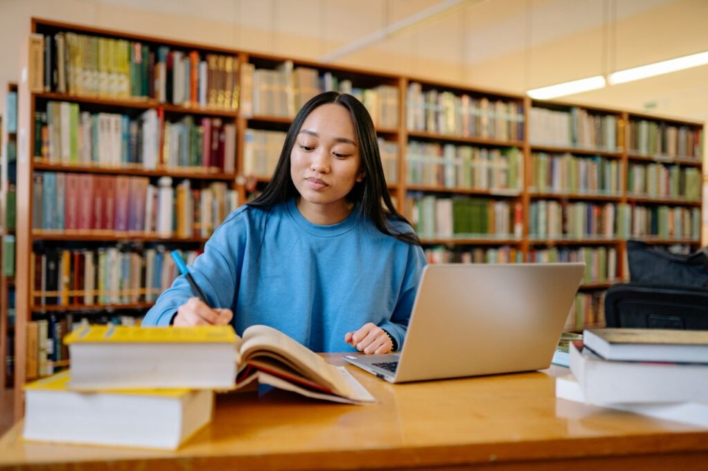 A student working at a laptop with books in a library, checking course details