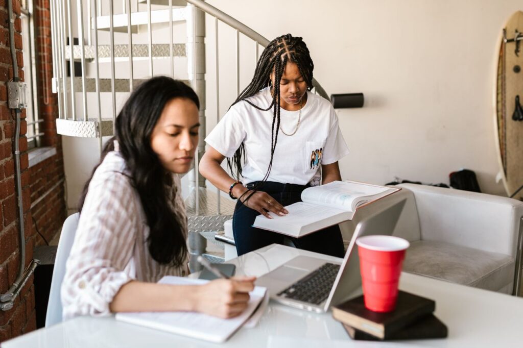 Two students looking at a laptop and a textbook together while researching options