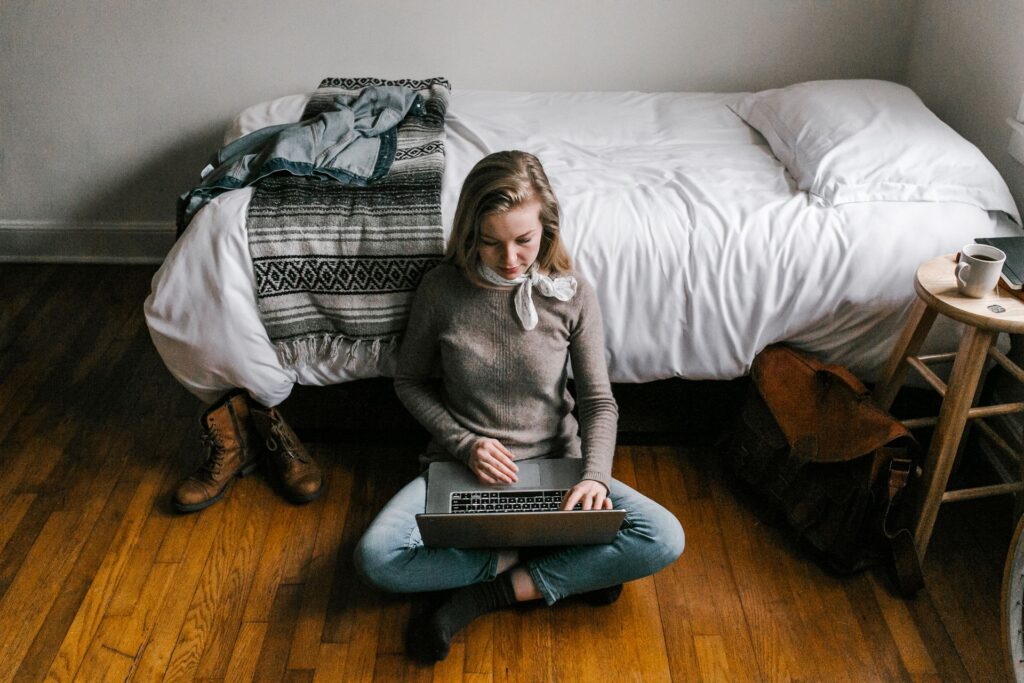 A student sitting on a bedroom floor searching for accommodation on a laptop