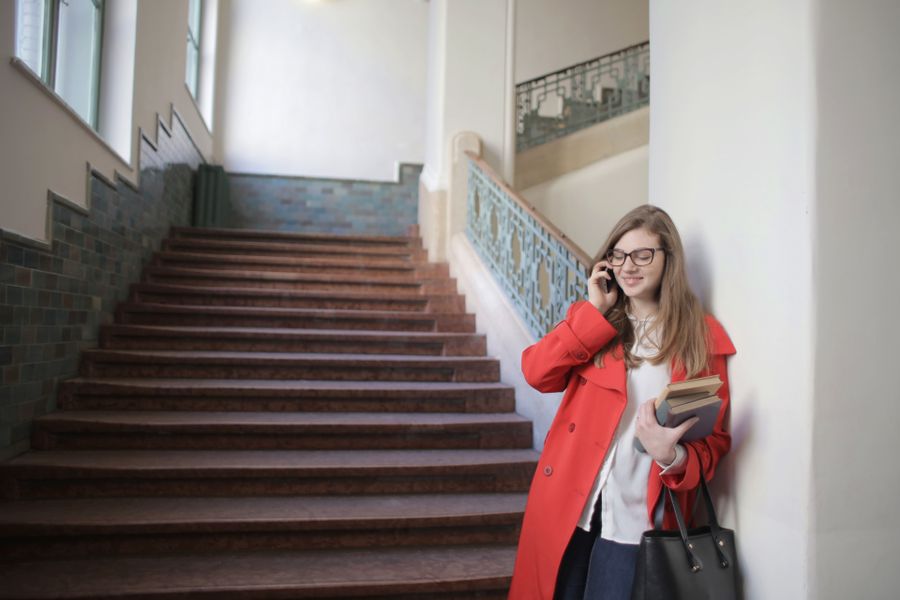 A student in an orange coat phones a university while holding her books on a staircase