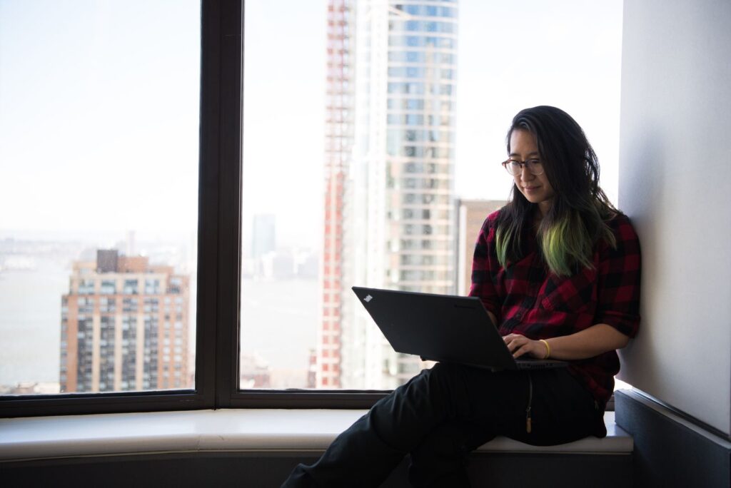 Graduate working on a laptop by a window overlooking a foreign city skyline while repaying a student loan abroad