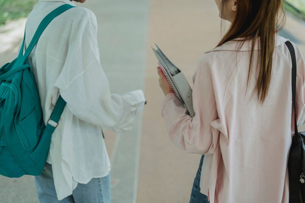 Two students with backpacks and notebooks walking together along a campus path, seen from behind.