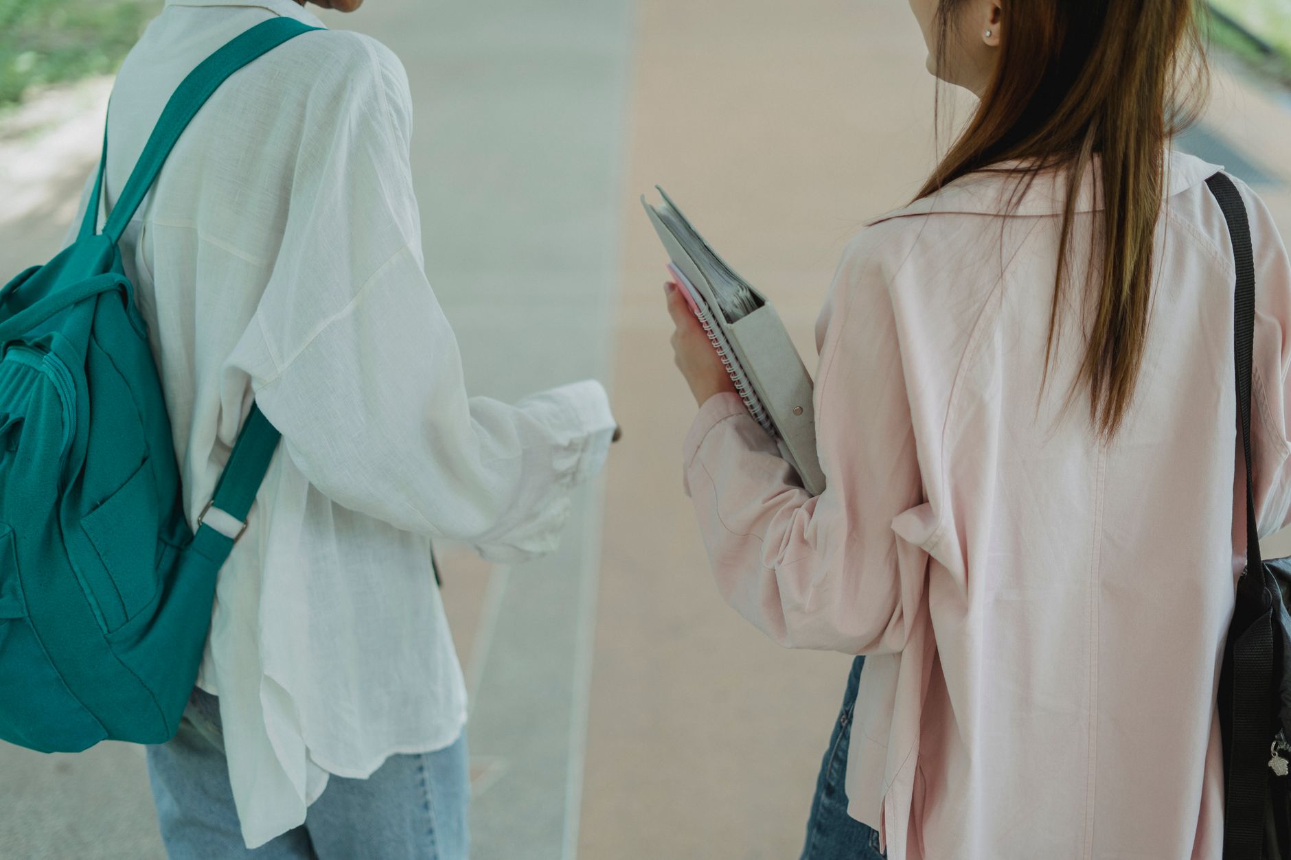 Two students with backpacks and notebooks walking together along a campus path, seen from behind.