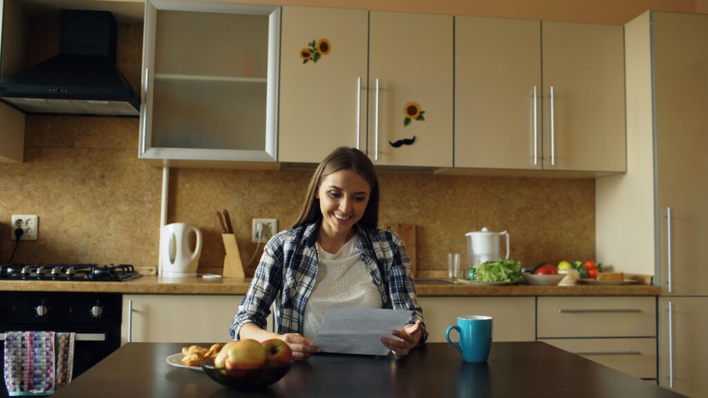Young woman reading a letter at a kitchen table with a laptop