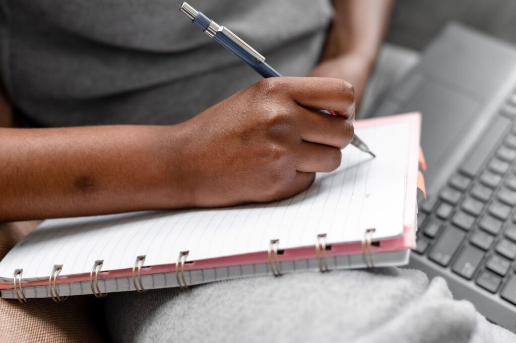 Student writing notes by hand in a spiral notebook next to a laptop