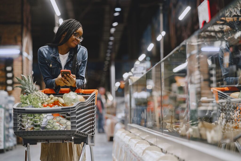 Young woman checking her phone beside a trolley of fresh food in a supermarket