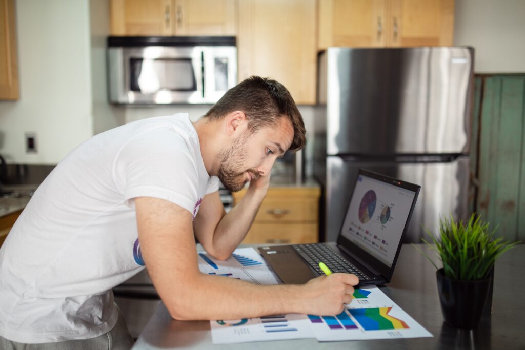 Man reviewing printed budget charts next to a laptop on a kitchen counter