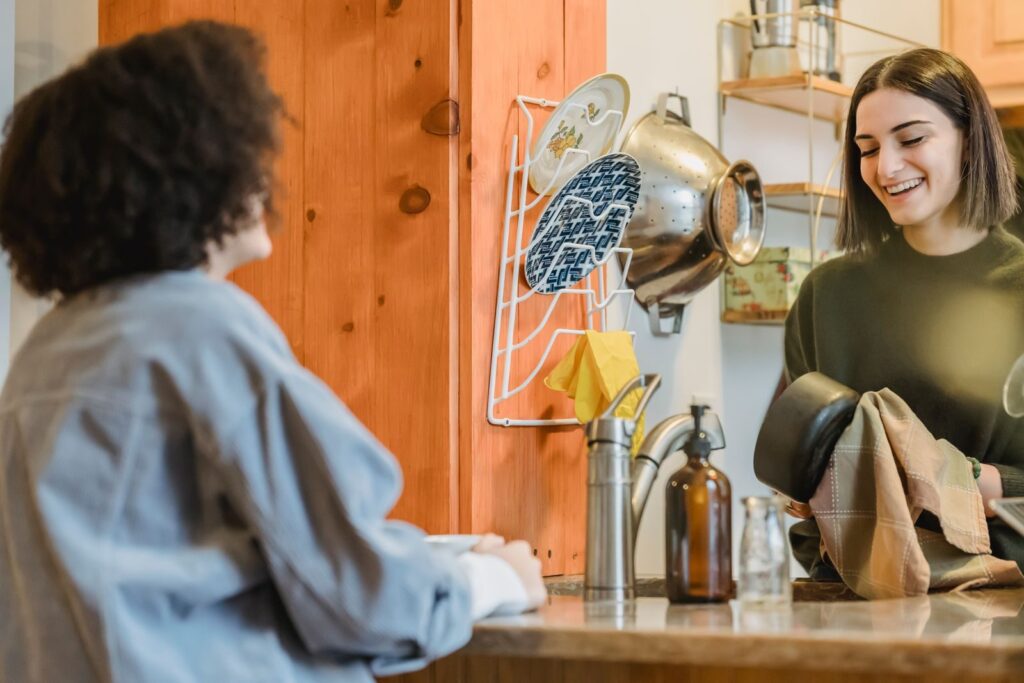 Two flatmates chatting across a shared kitchen counter while one dries the washing up