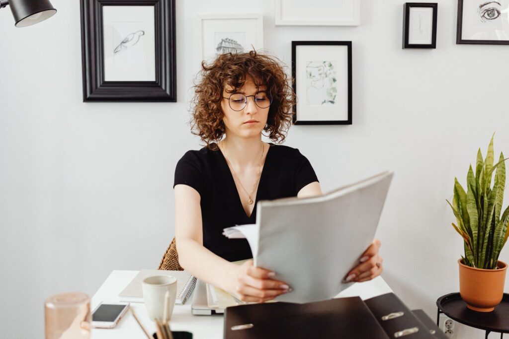 Young woman in glasses reading through printed documents in a folder at a desk