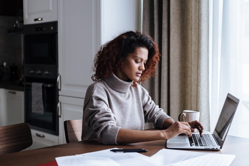 Young woman working through paperwork on a laptop at a kitchen table