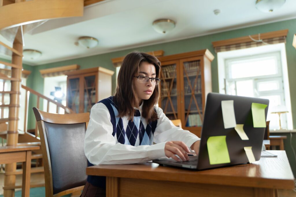 Student in glasses concentrating on redrafting work at a laptop in a library