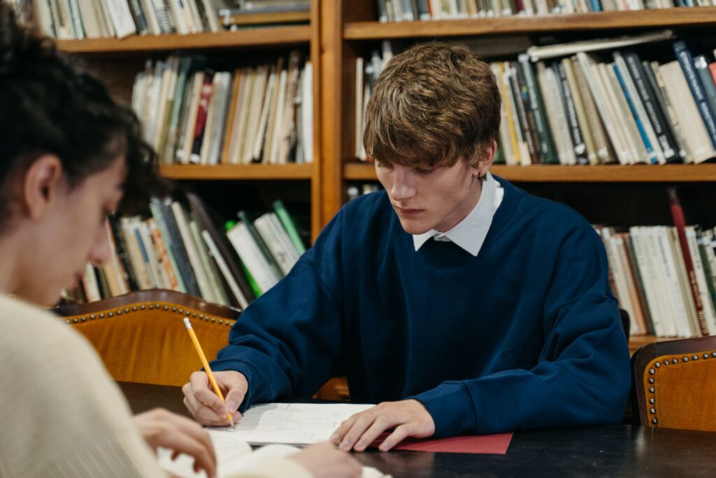 Two students working through notes at a library table surrounded by bookshelves
