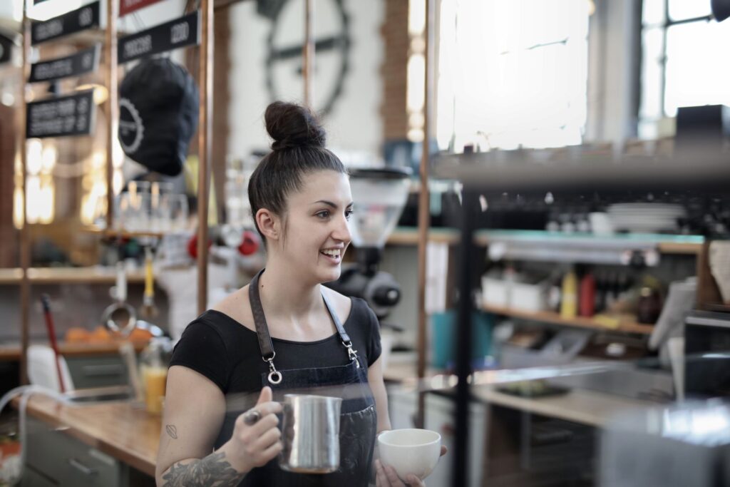 Young barista in an apron holding a milk jug and cup while working behind a cafe counter