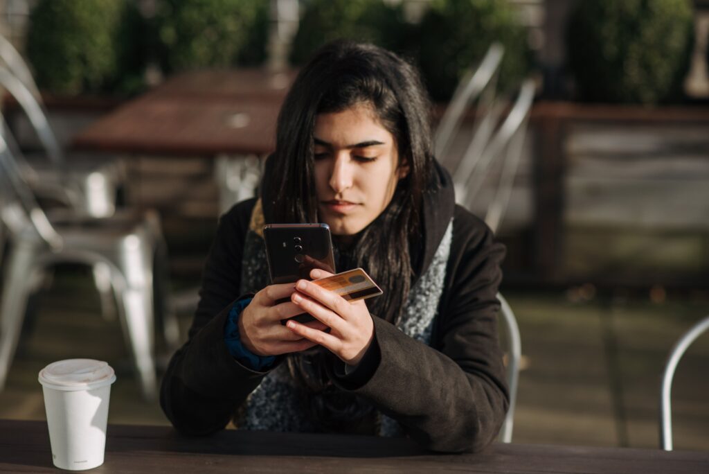Student at an outdoor cafe table holding a bank card while checking her phone