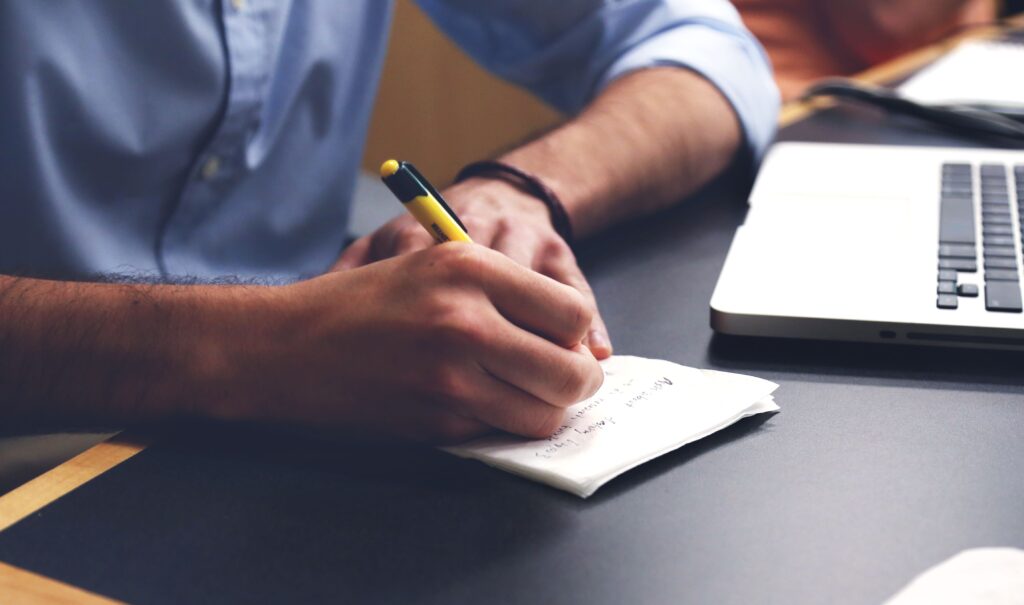 A person writing notes by hand next to a laptop on a desk