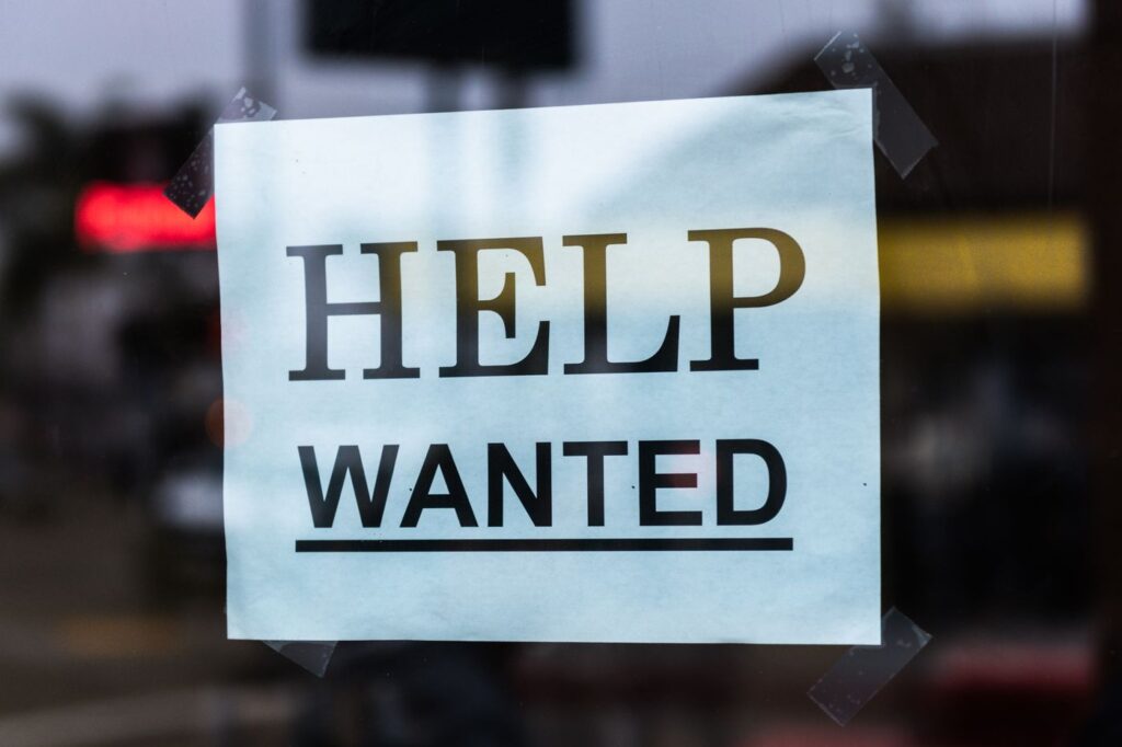 A Help Wanted sign taped inside a shop window, the kind of casual vacancy worth walking in to ask about