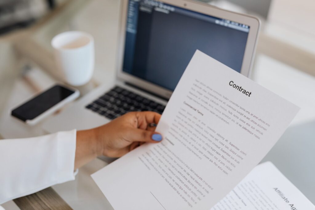 Person holding a printed contract beside a laptop, reading the terms before signing