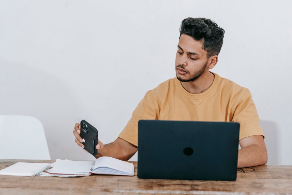 Young applicant at a desk checking his phone for updates next to a laptop and notebooks