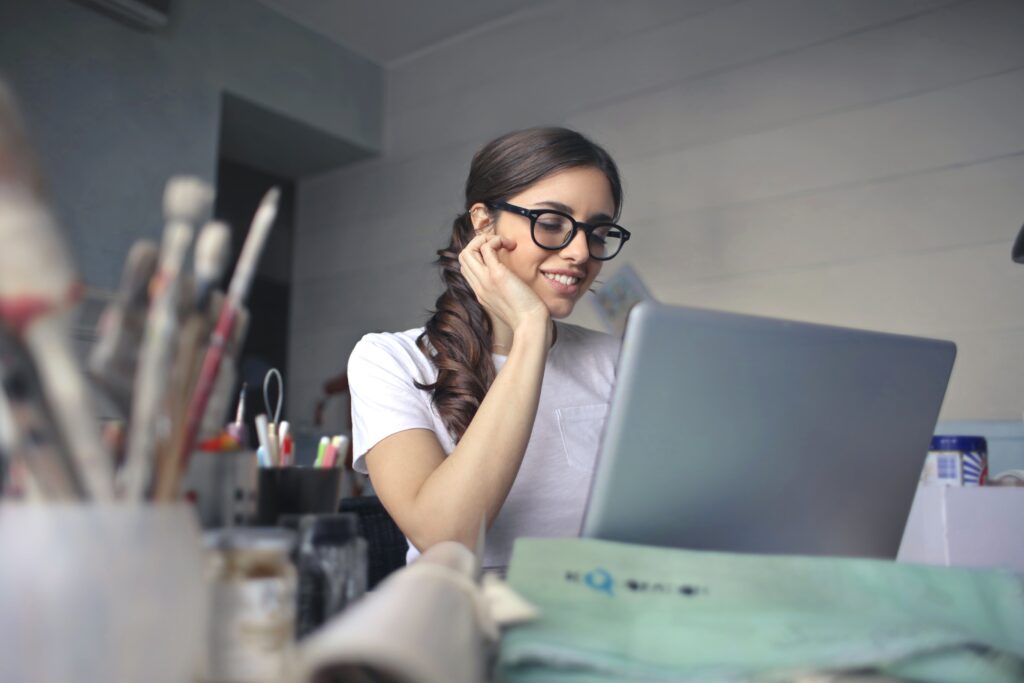 A student in glasses working at her laptop at a desk scattered with pens and brushes.