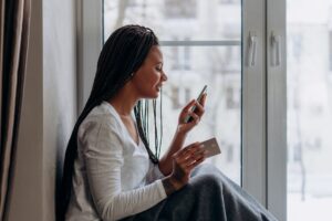 Woman sitting by the window shopping online with her phone and credit