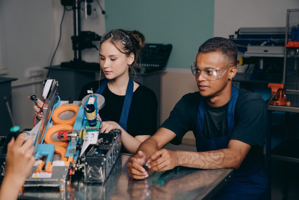 Two young apprentices in safety goggles working on a machine at a training workshop bench