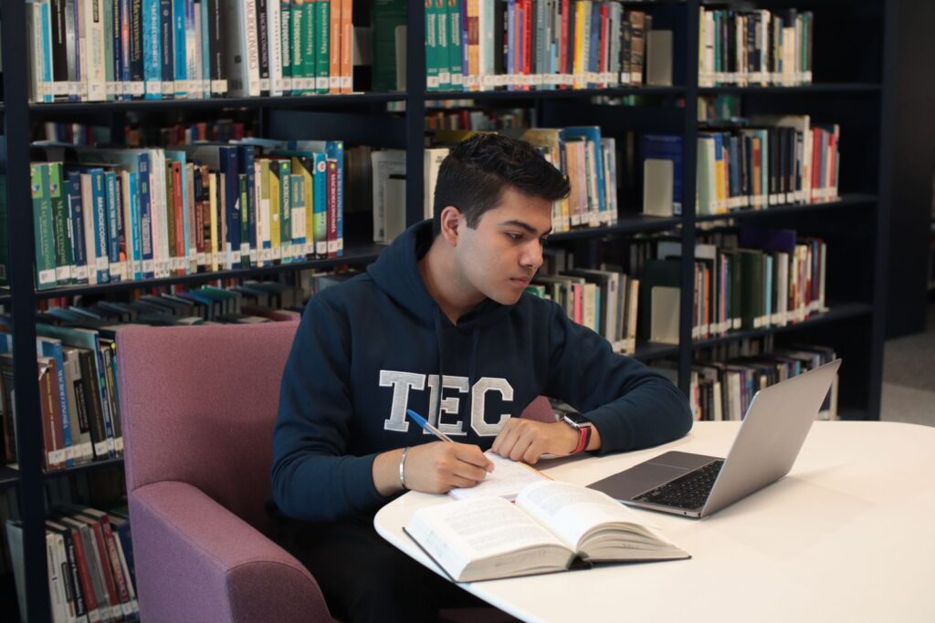 A student writing notes at a library desk with a laptop and a textbook open