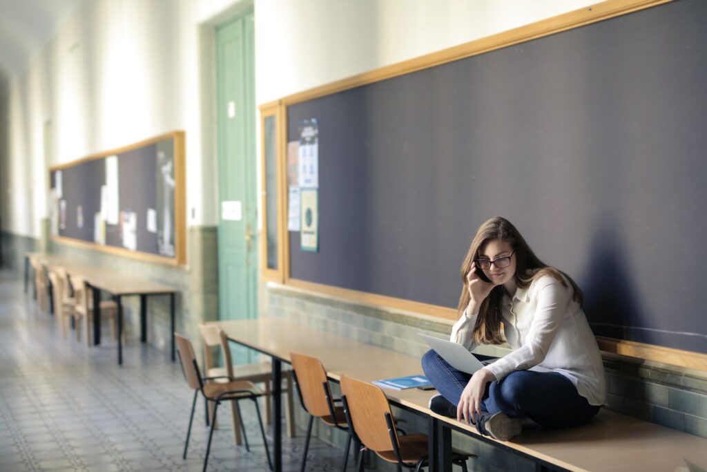 A student sits cross-legged on a bench in a university corridor, working on a laptop
