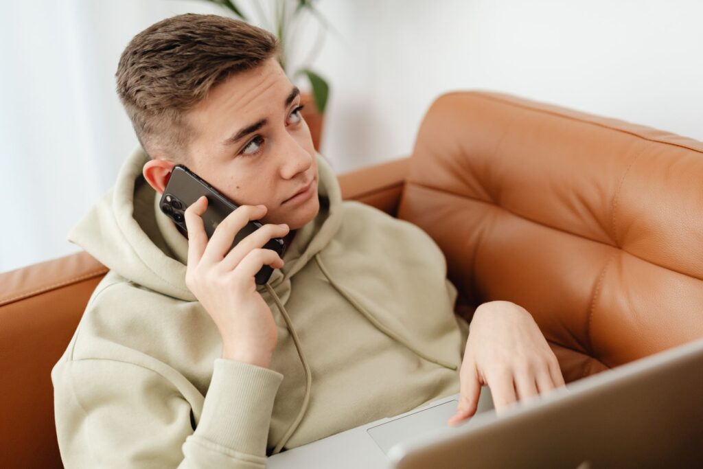 A teenager in a hoodie making a phone call from the sofa, laptop open on his lap