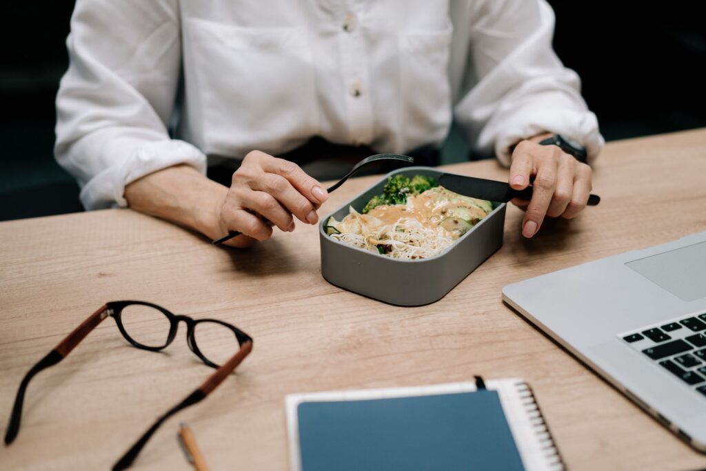A packed lunch in a metal tin being opened at an office desk beside a laptop and a pair of glasses